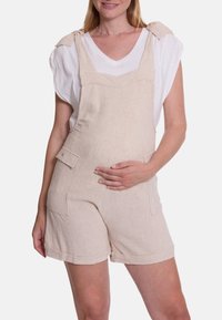 Beige linnen salopette met een witte v-hals t-shirt eronder. Heeft strikbanden, zijzakken en een omgerolde zoom. Glad oppervlak.
