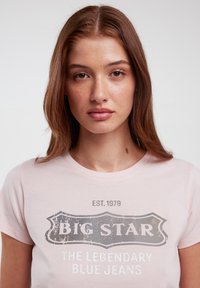 Šviesiai rožinė marškinėliai trumpomis rankovėmis su nusidėvėjusiu pilku logotipo spaudiniu, kuriame rašoma "BIG STAR EST. 1979 LEGENDINIAI MĖLYNI JANSAI."