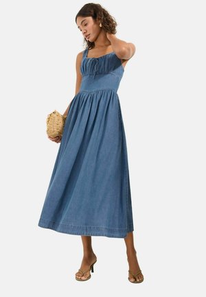 Kvinde iført en ærmeløs blå denimmidikjole, der holder en flettet clutch, står med den ene hånd ved halsen og har hælede sandaler på.