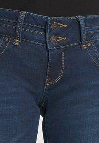 Primer plano de unos vaqueros de mezclilla de color azul oscuro con cierre de doble botón de metal y costuras amarillas en la cintura y los bolsillos.