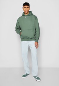 Sudadera verde con un bolsillo frontal y dobladillo acanalado, combinada con jeans de color azul claro y zapatillas verdes. Diseño sencillo, material de mezcla de algodón.