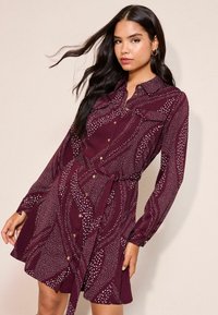 Bordeauxrotes Kleid mit langen Ärmeln, durchgehender Knopfleiste und tailliertem Gürtel. Verziert mit weißen abstrakten Mustern. Glattes Material mit einer figurbetonten Silhouette.