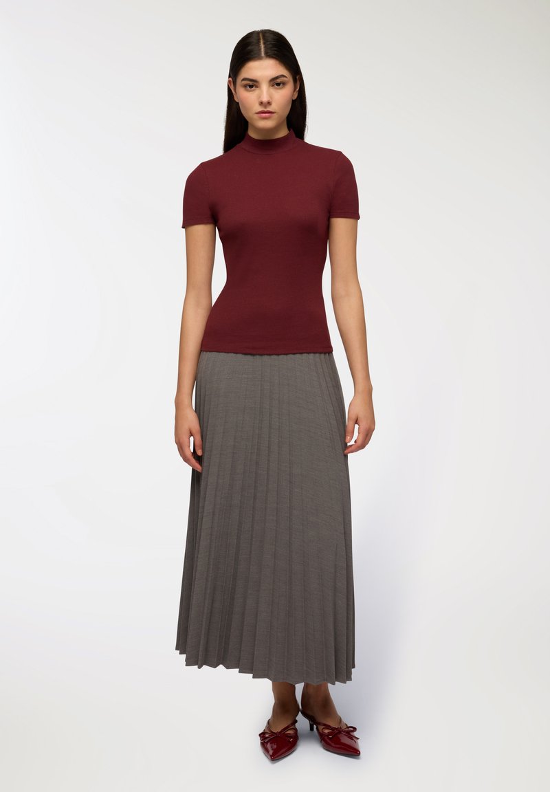 Top fitted a maniche corte color bordeaux abbinato a una gonna maxi grigia a pieghe. Il modello indossa scarpe con tacco rosse con un dettaglio a nodo sul davanti.