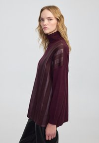 Kobieta z blond falującymi włosami ma na sobie fioletowy sweter z golfem ze skórzanymi paskami, w połączeniu z czarnymi spodniami, na tle jednolitego szarego tła.