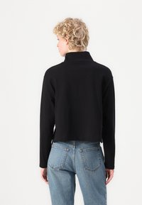 Černá zkrácená mikina s dlouhými rukávy a vysokým límcem, vyrobená z měkké tkaniny. Model má na sobě modré džíny s vysokým pasem.