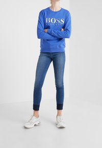 Modrý sweatshirt s kulatým výstřihem a bílým logem "BOSS HUGO BOSS", kombinovaný s úzkými džíny a bílými teniskami se šedými akcenty.