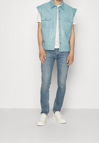 Person som bär en ljusblå ärmlös väst med knappar och fickor, vit t-shirt, blekta blå jeans och vita sneakers, stående mot en enfärgad bakgrund.