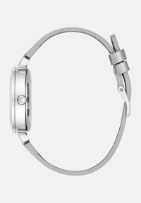 Sølvmetal ur med en rund kasse, slank rem og tekstureret hardware. Remmen er glat med en metallisk accent ved lukningen.