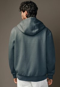 Mørk teal hoodie med et struktureret, vatteret mønster og en hætte med snor, der har ribbede manchetter og en båndet kant.