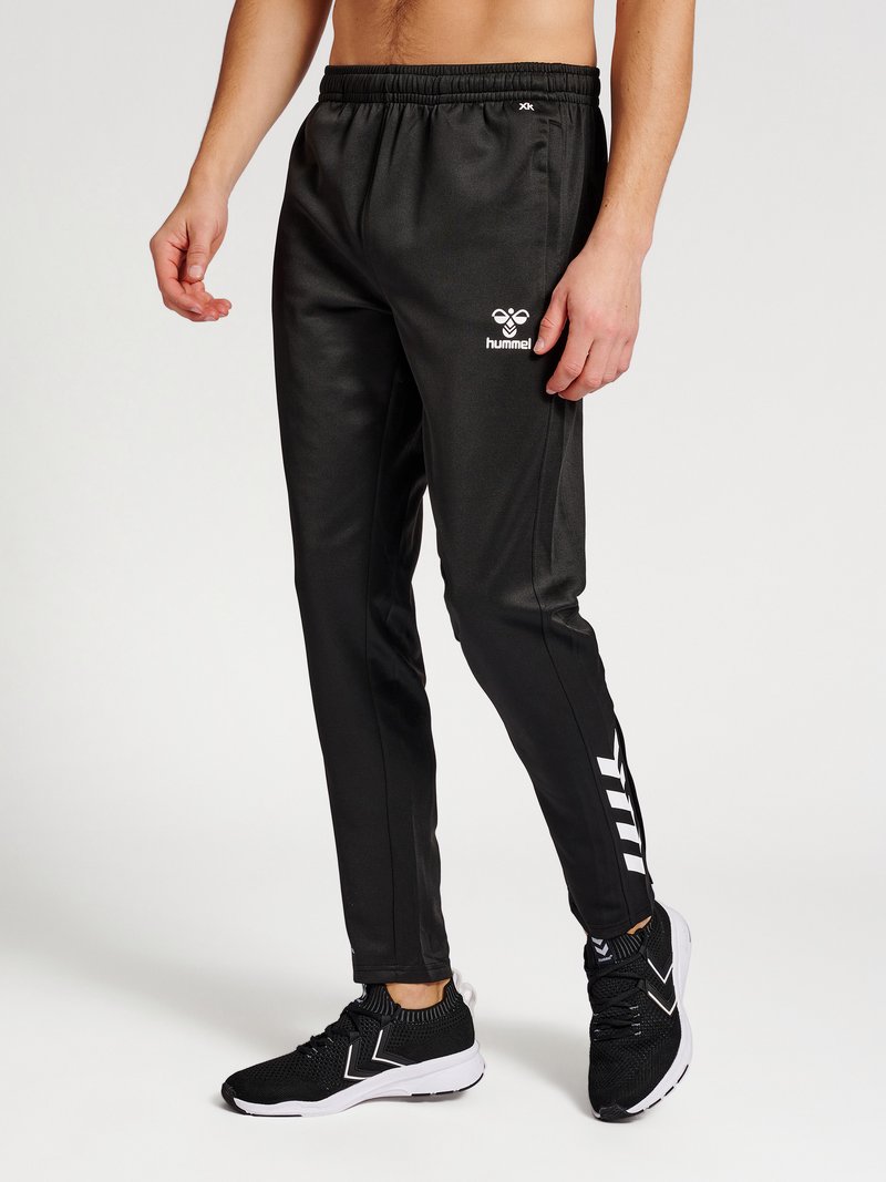 Uomo che indossa pantaloni da tuta neri Hummel e scarpe sportive nere, in piedi davanti a uno sfondo semplice.