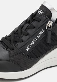 Baskets Michael Kors noires avec détails texturés, lacets noirs, fermeture éclair latérale, logo blanc et semelle blanche avec rainures noires.