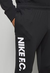 Černé sportovní kalhoty vyrobené z hladké látky s elastickým pasem. Na bocích se nachází velké bílé logo "NIKE F.C.".