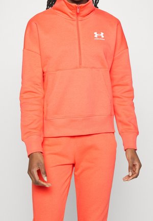 Personne portant un sweat à demi-fermeture éclair Under Armour corail vif et un pantalon assorti, les mains détendues le long du corps devant un fond uni.