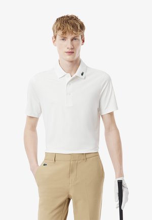 Hvid polo skjorte med krave, der har et sort broderet logo på kraven. Båret med beige bukser og en golfhandske.