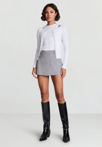 Cardigan blanc à manches longues côtelé avec boutons, associé à une mini-jupe grise et des bottes en cuir noires montant jusqu'au genou. Design et textures simples.