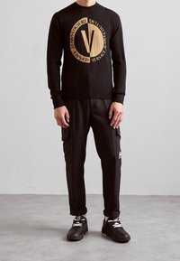Pull en maille noire avec un grand logo doré au centre, assorti à un pantalon cargo noir et des chaussures de sport noires avec des lacets blancs.
