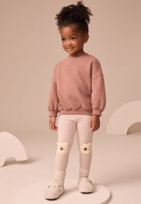 Svetloružová mikina s nafúknutými rukávmi, v kombinácii s krémovými pruhovanými legínami s medvedíkovými aplikáciami na kolenách a mäkkými béžovými papučami.