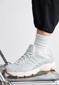 Vaaleanharmaa urheilukenkä, jossa on mokkaa ja verkkomateriaaleja. Kenkässä on kolme rinnakkaista raitaa, valkoinen pohja ja kumipohja. Kenkä on yhdistetty raidoitettuun sukkaan.