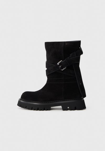 Schwarze Wildlederstiefel bis zur Wade mit dicker Profilsohle und überkreuzten verstellbaren Riemen mit silbernen Schnallen auf einem einfarbigen Hintergrund.