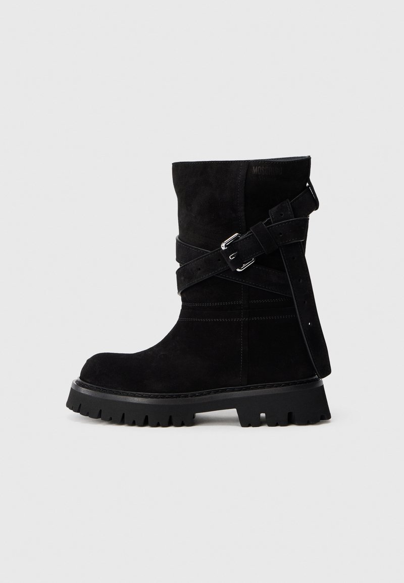 Bota de ante negra hasta la mitad de la pantorrilla con suela gruesa y estriada, y correas ajustables entrecruzadas con hebillas plateadas sobre un fondo liso.