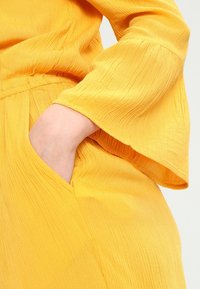 Gul klänning med texturerad tyg; har klockärmar, en figursydd midja och sidofickor. Materialet har synliga vertikala linjer.