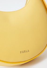 Žuta kožna torbica s zaobljenim dizajnom, glatkom teksturom i utisnutim logotipom "FURLA" s prednje strane. Jednostavna i elegantna izrada.