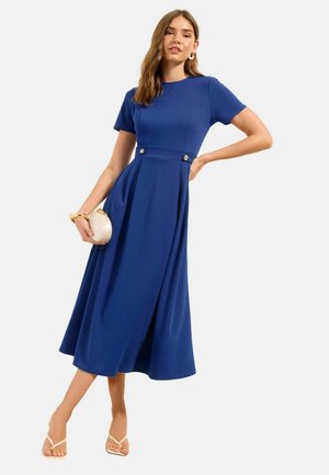 Frau in einem blauen Midikleid mit kurzen Ärmeln, die eine beige Clutch hält und beige Sandalen trägt, stehend mit einer Hand in der Hüfte.