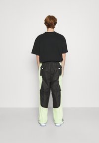 Černé oversized tričko a černé kalhoty s neonově zelenými síťovinovými bočními panely, obsahující cargo kapsy a uvolněný střih, viděné zezadu.