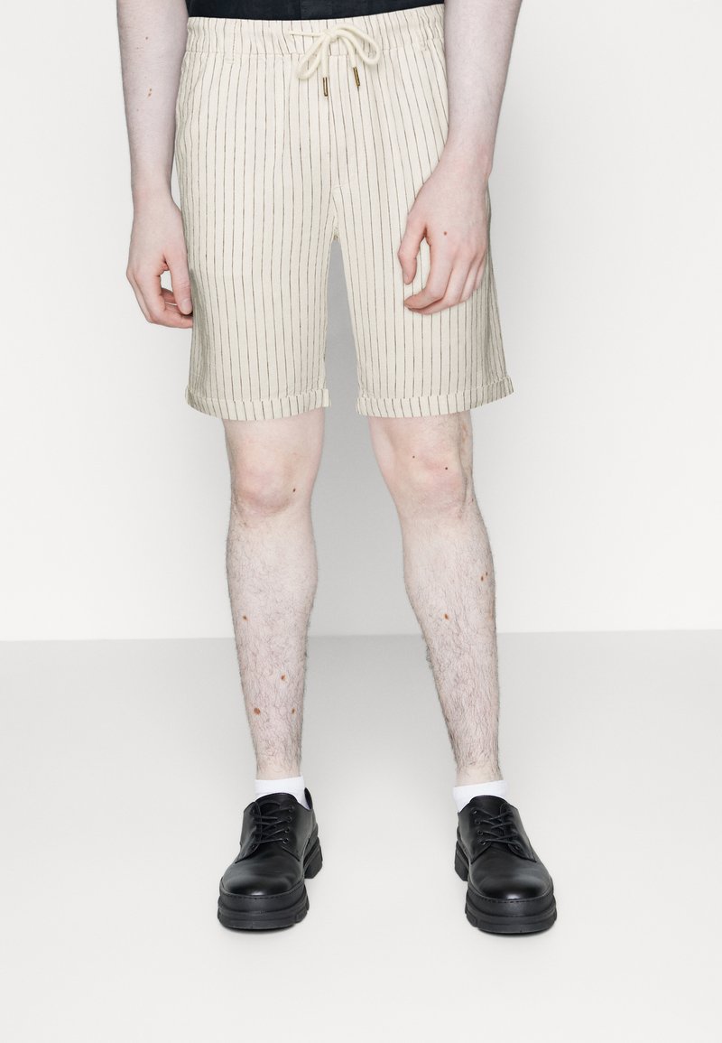 Shorts beiges à rayures avec une ceinture élastique et un cordon de serrage. Texture lisse, ourlet roulé, portés avec des chaussures noires et des chaussettes blanches.