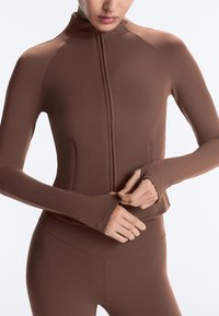 Frau trägt eng anliegende braune Zip-Up-Sportjacke und passende Leggings, richtet den unteren Rand der Jacke mit beiden Händen.