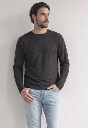 Homme aux cheveux foncés et à la barbe, portant une chemise noire à manches longues et un jean bleu clair, debout devant un fond uni clair.