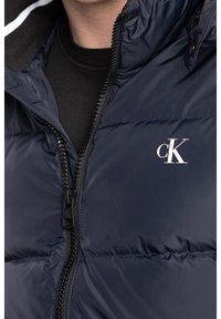 Mörkblå puffjacka med dragkedja, med hög krage, sidofickor och en liten vit logotyp på bröstet. Slät textur.