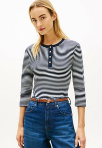 Kobieta ubrana w granatowo-białą koszulę w paski z długim rękawem, ozdobioną złotymi guzikami, oraz wysokie niebieskie dżinsy z brązowym paskiem.