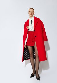 Roter Wollmantel mit großen schwarzen Knöpfen, kombiniert mit einem strukturierten roten Miniröckchen. Weißes Bluse mit hohem Kragen und transparente schwarze Strumpfhosen zu spitzen schwarzen Schuhen.