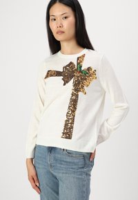 Witte trui met een gouden paillettenstrikdesign en groene accenten. Gemaakt van zacht materiaal, met een ronde hals en lange mouwen.