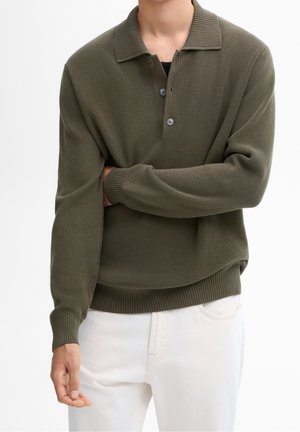 Mężczyzna w oliwkowo-zielonym prążkowanym, dzianinowym swetrze polo z długim rękawem i dwoma guzikami, w połączeniu z białymi spodniami, częściowo skrzyżowanymi ramionami.