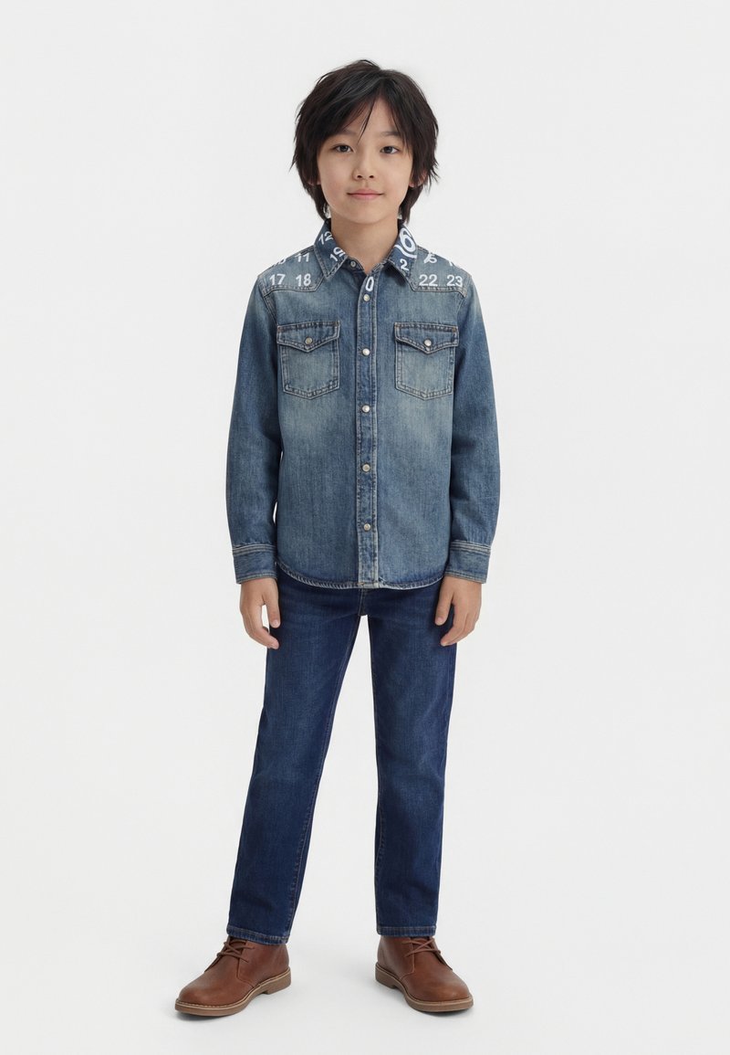 Αγόρι όρθιο, φοράει μια μπλε τζιν πουκάμισο με κουμπιά και τυπωμένους αριθμούς στους ώμους, σκούρα τζιν και καφέ δερμάτινα παπούτσια.