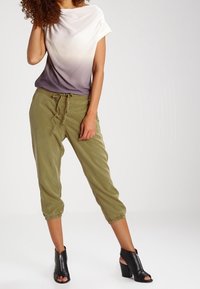Mujer de pie lleva una blusa en degradado de blanco a gris, pantalones cortos de color verde oliva con cordón y botines de tacón abierto negros.