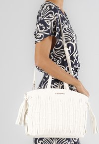 Valkoinen käsilaukku, jossa on kudontakuosi, tekstiilimuotoilun yksityiskohtia ja tupsu, jonka kantaa henkilö, joka on pukeutunut tummansiniseen ja valkoiseen kuosilliseen paitaan.