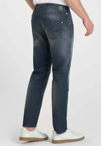 Jeans de mezclilla azul oscuro con corte ajustado; cuentan con bolsillos traseros y una etiqueta de cuero en la cintura. Se combinan con zapatillas blancas y una camiseta casual.