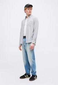 Jeune homme portant une casquette noire, une chemise à boutons gris clair par-dessus un t-shirt blanc, un jean bleu clair et des baskets noires à rayures beige, debout de profil.
