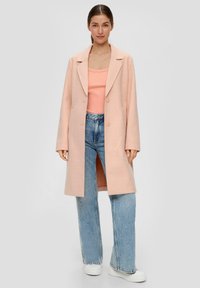 Różowy, oversize'owy płaszcz wełniany z klapami i podwójnymi guzikami, zestawiony z prążkowanym różowym topem i jasnoniebieskimi spodniami typu wide-leg. Białe sneakersy.