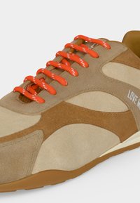 Gros plan d'une basket en daim beige et marron avec des lacets orange vif et un texte blanc partiel sur le côté.