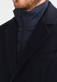 Navy wool coat with a high collar, featuring a quilted inner layer and zip closure. Textured fabric and subtle stitching detail are visible.