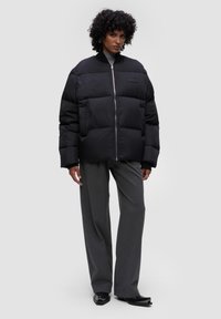 Czarna kurtka puchowa z wysokim kołnierzem, przednim zamkiem błyskawicznym, dużymi puchowymi sekcjami i bocznymi kieszeniami, noszona z szarymi spodniami o szerokich nogawkach i czarnymi butami.