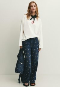 Jonge vrouw draagt een witte oversized sweatshirt, donkerblauwe spijkerbroek met zilveren oogjes, houdt een bijpassende spijkerjas met oogjes vast en draagt zwarte sandalen.