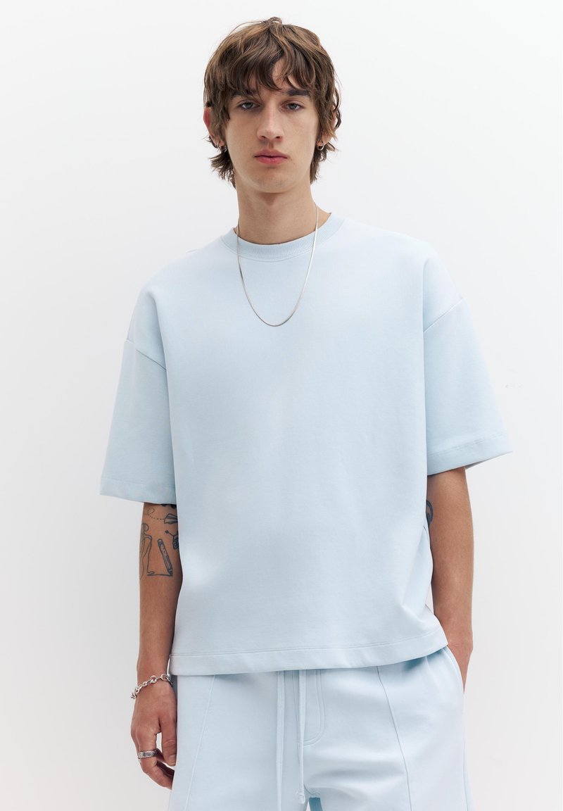 Jeune homme aux cheveux bruns mi-longs portant un t-shirt oversize bleu clair et un pantalon assorti, accessoirisé avec un collier chaîne, un bracelet et des bagues.
