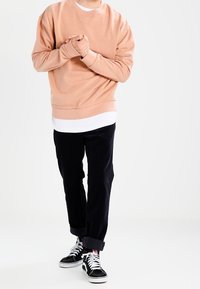 Personne portant un sweat à capuche pêche, un t-shirt blanc en dessous, un pantalon noir retroussé et des baskets montantes noires, se tenant debout avec les mains jointes.