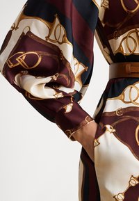 Kvinnehånd i lommen på mønstret satengkjole med ridderinspirert design i brunt, gull, krem og svart, med belte.