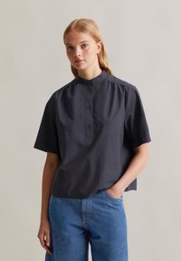 Maglietta grigio scuro a maniche corte con colletto rialzato, chiusura frontale a bottoni e leggero arricciamento sulle spalle, abbinata a jeans in denim blu.